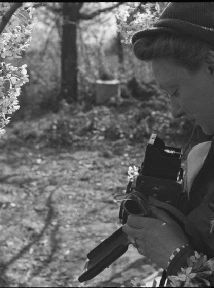 Photographie en noir et blanc de Germaine Kanova, en uniforme de correspondante de guerre, tenant un appareil photo au milieu des fleurs.