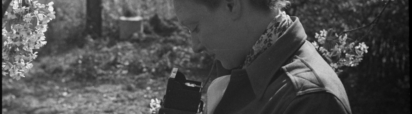 Photographie en noir et blanc de Germaine Kanova, en uniforme de correspondante de guerre, tenant un appareil photo au milieu des fleurs.