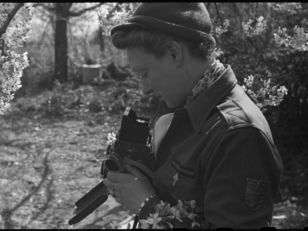 Photographie en noir et blanc de Germaine Kanova, en uniforme de correspondante de guerre, tenant un appareil photo au milieu des fleurs.