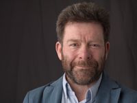 Portrait en intérieur d'un homme d'une cinquantaine d'années, cheveux et barbe grisonnants courts, veste bleu marine sur chemise, souriant légèrement, fond sombre uni.