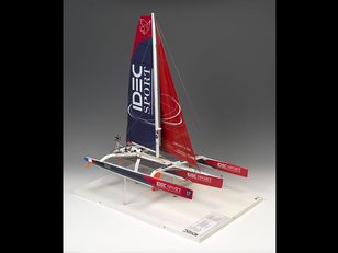 Model sailboat with a red and white sail that has "IDEC SPORT" written on it a white hull, and a red and white striped flag on the top of the mast. The sailboat is mounted on a white base 