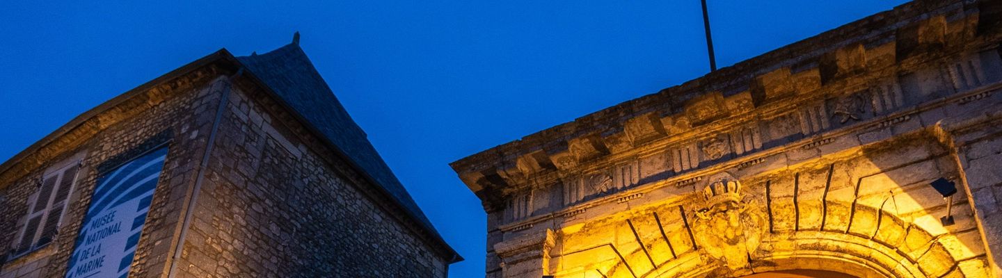 Façade illuminée du musée national de la Marine à Rochefort à la tombée de la nuit, avec son portail d'entrée en pierre.