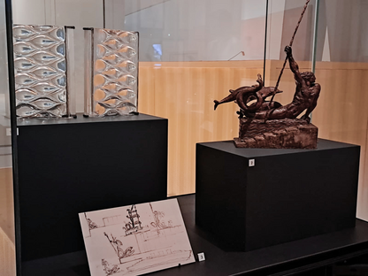 Vitrine d’exposition avec maquette sculptée du Génie de la mer, dessins préparatoires du paquebot Normandie et éléments de luminaires en verre de René Lalique présentés sur socles noirs.