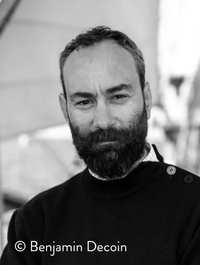 Portrait en noir et blanc d’un homme barbu debout à bord d’un voilier, avec les voiles floues en arrière-plan.