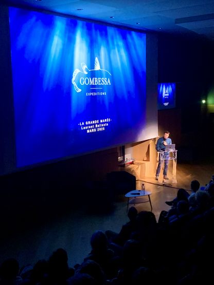 Laurent Ballesta intervenant au musée national de la Marine, devant un écran projetant le visuel « Gombessa Expeditions – La Grande Marée – mars 2025 » aux couleurs des profondeurs marines.