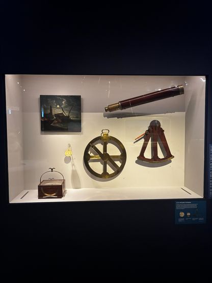 Vitrine du Mucem présentant quatre instruments de navigation anciens : sextant, lunette, horloge marine, roue de gouvernail. Et un tableau
