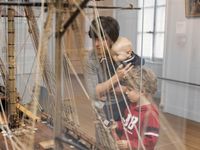 Maman tenant un bébé dans les bras accompagnée d'un enfant plus grand regardant une maquette