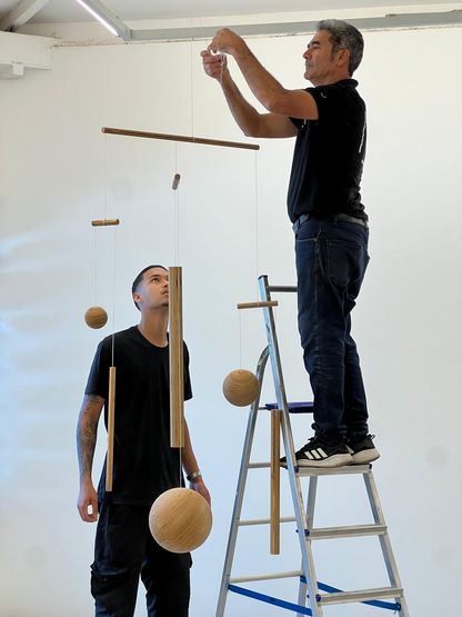 Deux hommes installent une œuvre suspendue composée d’éléments en bois sphériques et cylindriques dans un atelier, l’un sur une échelle ajustant la structure, l’autre observant depuis le sol.
