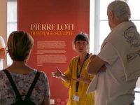 La commissaire de l'exposition anime une visite guidée de l’exposition "Pierre Loti, hommage sculpté", en interaction avec un petit groupe de visiteurs.