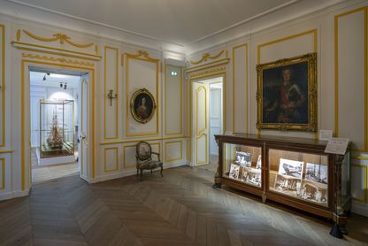 Salon de compagnie du parcours permanent rénové du Musée national de la Marine à Rochefort, avec boiseries blanches et dorées, tableau ancien dans un cadre doré, vitrine d'exposition ornée en premier plan et vue en enfilade vers une salle voisine.