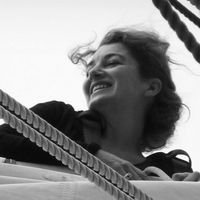 Portrait en noir et blanc d’une femme souriante regardant vers le large, appuyée contre le gréement d’un voilier.