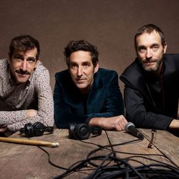 Photo des trois artistes accoudés sur une table présentant des accessoires de musique : un casque, un micro, des baguettes de batterie, un cable.