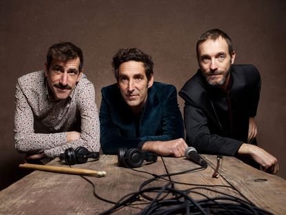 Photo des trois artistes accoudés sur une table présentant des accessoires de musique : un casque, un micro, des baguettes de batterie, un cable.