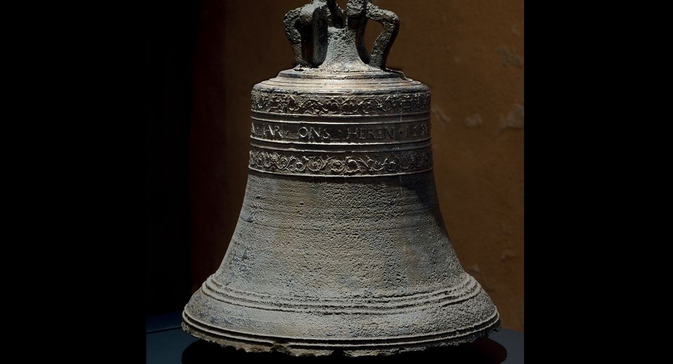 A photo realistic image of an ancient bronze bell on a black pedestal with intricate designs and patterns on its surface. The bell has a handle on top. 